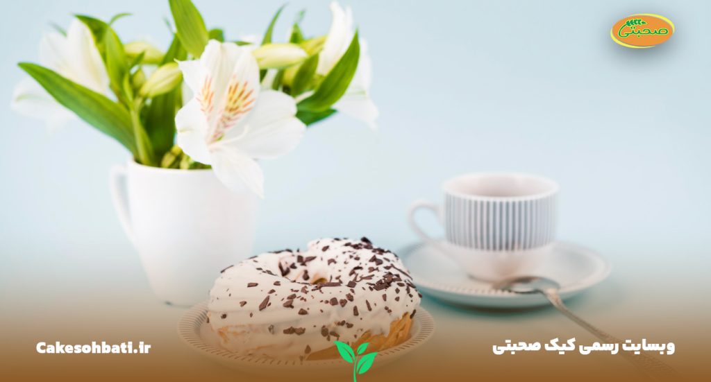 تصویری از یک کیک وانیلی در کنار یک فنجان و در کنار آنها یک دسته گل وانیل درون لیوانی قرار داده شده است.