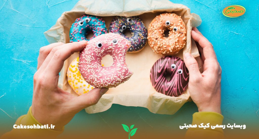 چند عدد دونات با طراحی های رنگین و متفاوت درون یک جعبه و شخصی که یک دونات خورده شده را دست دارد.