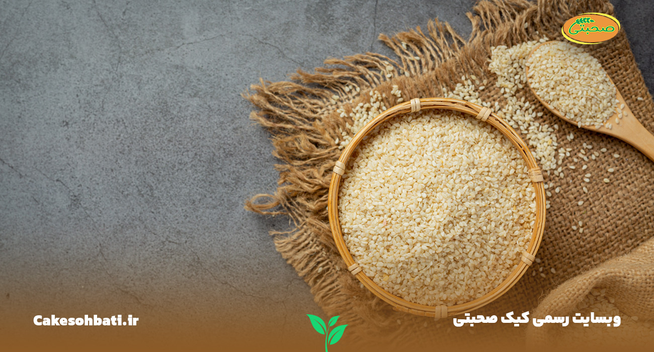 نمایی بالا از تصویری با پس زمینه تیره که بر روی یک عدد پارچه قهوه ای وجود دارد و بر روی آن یک کاسه بزرگ کنجد که در کنار آن دانه های کنجد پراکنده هستند.