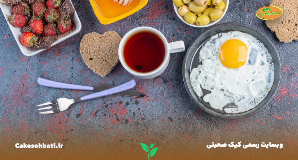 تصویری از نمای بالا که چند نوع صبحانه فوری و کامل را نشان می‌دهد. یک لیوان چای، یک قاشق و چنگال، یک تخم مرغ آماده شده، یک ظرف زیتون، یک تکه نان جو با طرح قلب و یک ظرف شکلات و توت فرنگی در تصویری دیده می‌شود.