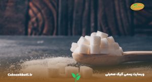 تصویری از نمای کناری از یک میز تیره رنگ که بر روی آن مقداری شکر و قند پاشیده شده است و قاشقی که در آن چند حبه قند سنتزی دیده می‌شود.